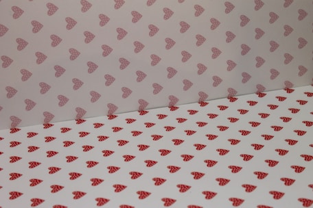 A pattern of heart shapes covers a surface and wall. The hearts are red with polka dots on a white background. The upper section has a lighter shade with similar heart patterns.