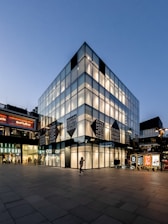 A modern, multi-story glass building with reflective panels, illuminated from within during dusk. The lower floors display large signage reading 'Dover Street Market Beijing.' People are seen walking around, and nearby are brightly lit advertisement boards, enhancing the urban atmosphere.