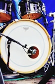 A close-up view of a drum set featuring a bass drum with a microphone placed in front of it. The drumhead has a logo and is bordered by a yellow rim. Additional drum components are visible in the background with metallic hardware and a blue backdrop.