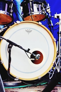 A close-up view of a drum set featuring a bass drum with a microphone placed in front of it. The drumhead has a logo and is bordered by a yellow rim. Additional drum components are visible in the background with metallic hardware and a blue backdrop.