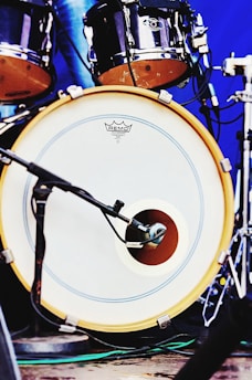 A close-up view of a drum set featuring a bass drum with a microphone placed in front of it. The drumhead has a logo and is bordered by a yellow rim. Additional drum components are visible in the background with metallic hardware and a blue backdrop.