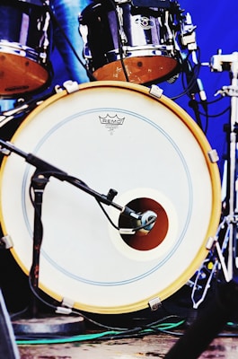 A close-up view of a drum set featuring a bass drum with a microphone placed in front of it. The drumhead has a logo and is bordered by a yellow rim. Additional drum components are visible in the background with metallic hardware and a blue backdrop.