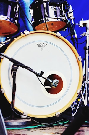 A close-up view of a drum set featuring a bass drum with a microphone placed in front of it. The drumhead has a logo and is bordered by a yellow rim. Additional drum components are visible in the background with metallic hardware and a blue backdrop.