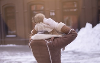 A person is pictured from behind wearing a brown winter coat with white trim, a beige knitted hat with a fur pom-pom, and white mittens. The individual appears to be adjusting or holding their hat. The background consists of a wintry urban setting with snow on the ground and a building with dark windows.