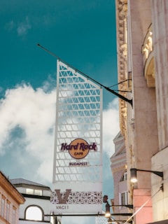 A street-level view features a prominent sign for the Hard Rock Cafe in Budapest. The sign is attached to a modern, triangular lattice structure with a clear blue sky and a few clouds in the background. Surrounding the sign are elements of classic architecture from nearby buildings.