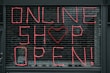 Large letters formed in pink string spell out 'ONLINE SHOP OPEN!' on a black metal grid. A heart shape replaces the letter 'O' in the word 'SHOP'. The scene seems to depict a storefront or window display.