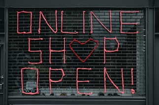Large letters formed in pink string spell out 'ONLINE SHOP OPEN!' on a black metal grid. A heart shape replaces the letter 'O' in the word 'SHOP'. The scene seems to depict a storefront or window display.