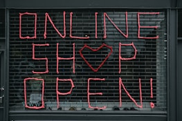 Large letters formed in pink string spell out 'ONLINE SHOP OPEN!' on a black metal grid. A heart shape replaces the letter 'O' in the word 'SHOP'. The scene seems to depict a storefront or window display.