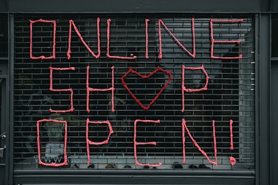 Large letters formed in pink string spell out 'ONLINE SHOP OPEN!' on a black metal grid. A heart shape replaces the letter 'O' in the word 'SHOP'. The scene seems to depict a storefront or window display.