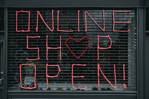 Large letters formed in pink string spell out 'ONLINE SHOP OPEN!' on a black metal grid. A heart shape replaces the letter 'O' in the word 'SHOP'. The scene seems to depict a storefront or window display.