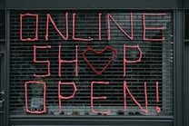 Large letters formed in pink string spell out 'ONLINE SHOP OPEN!' on a black metal grid. A heart shape replaces the letter 'O' in the word 'SHOP'. The scene seems to depict a storefront or window display.
