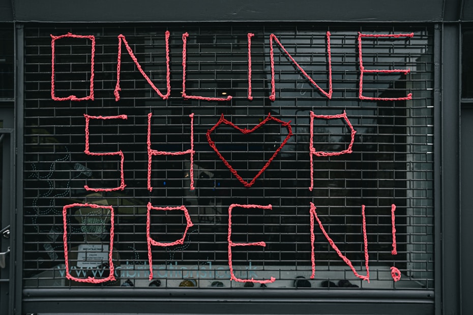 Large letters formed in pink string spell out 'ONLINE SHOP OPEN!' on a black metal grid. A heart shape replaces the letter 'O' in the word 'SHOP'. The scene seems to depict a storefront or window display.