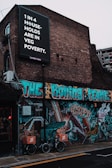 A brick building is adorned with vibrant street art featuring bold, colorful graffiti. Above, a large black and white sign with text highlights an issue regarding 'veg poverty' affecting one in four households. Two bicycles with cargo boxes labeled 'Yard Sale Pizza' are parked in front of the mural, which showcases various abstract designs and figures.