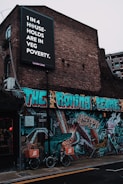 A brick building is adorned with vibrant street art featuring bold, colorful graffiti. Above, a large black and white sign with text highlights an issue regarding 'veg poverty' affecting one in four households. Two bicycles with cargo boxes labeled 'Yard Sale Pizza' are parked in front of the mural, which showcases various abstract designs and figures.