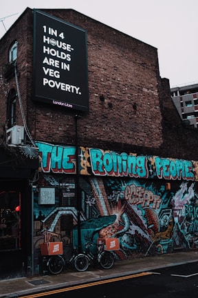 A brick building is adorned with vibrant street art featuring bold, colorful graffiti. Above, a large black and white sign with text highlights an issue regarding 'veg poverty' affecting one in four households. Two bicycles with cargo boxes labeled 'Yard Sale Pizza' are parked in front of the mural, which showcases various abstract designs and figures.