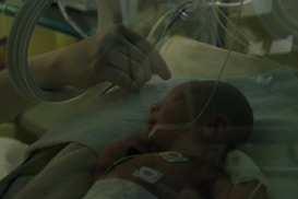 A newborn baby lies in an incubator, covered with a soft blanket and attached to medical monitoring equipment. A gentle hand reaches in to touch or comfort the infant, creating a sense of care and protection.