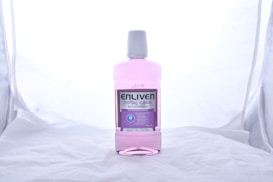 A bottle of Enliven Total Care Mouthwash is centered against a plain, light-colored background. The mouthwash is pink and packaged in a clear plastic bottle with a white cap. The label is predominantly purple and white, providing information about the product, such as its 6 benefits.