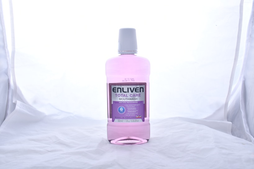 A bottle of Enliven Total Care Mouthwash is centered against a plain, light-colored background. The mouthwash is pink and packaged in a clear plastic bottle with a white cap. The label is predominantly purple and white, providing information about the product, such as its 6 benefits.