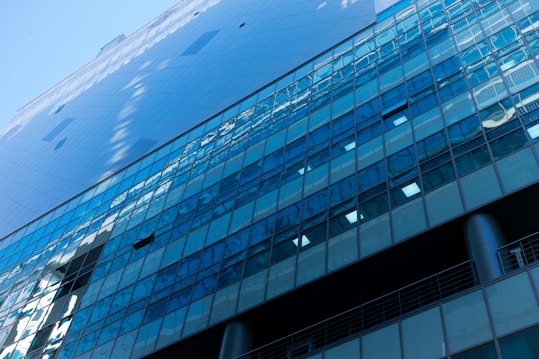 A high-rise building facade features numerous reflective glass windows, with a modern, sleek design. The architecture includes vertical metallic columns and a pattern of windows that capture the sky and surrounding structures. The angle of the photograph emphasizes the height and geometric patterns of the building.