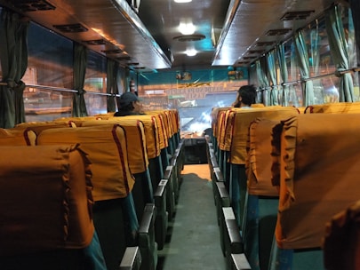 The interior of a bus is depicted with rows of seats covered in orange-tinted seat covers. There are two people sitting at the front of the bus, facing forward. The bus has windows adorned with curtains, and there is a view of lights and signs outside through the windows.
