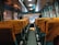The interior of a bus is depicted with rows of seats covered in orange-tinted seat covers. There are two people sitting at the front of the bus, facing forward. The bus has windows adorned with curtains, and there is a view of lights and signs outside through the windows.