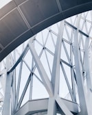 Abstract architectural structure featuring intersecting metal beams and large glass panels forming geometric patterns. The design appears modern and intricate, showcasing a blend of curves and straight lines.