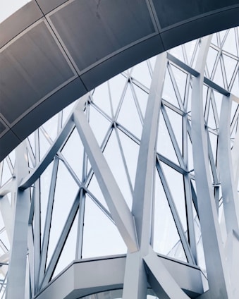 Abstract architectural structure featuring intersecting metal beams and large glass panels forming geometric patterns. The design appears modern and intricate, showcasing a blend of curves and straight lines.