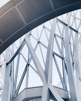 Abstract architectural structure featuring intersecting metal beams and large glass panels forming geometric patterns. The design appears modern and intricate, showcasing a blend of curves and straight lines.