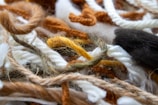 A close-up view of various colored yarns and threads intertwined in a chaotic bundle. The fibers include a mix of textures and thicknesses, with soft and fluffy appearances. The colors range from whites and browns to oranges and blacks.