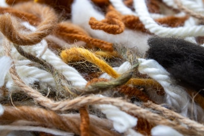 A close-up view of various colored yarns and threads intertwined in a chaotic bundle. The fibers include a mix of textures and thicknesses, with soft and fluffy appearances. The colors range from whites and browns to oranges and blacks.