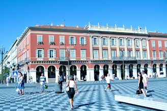 A large plaza with a checkered black and white pattern on the ground is bordered by a historic building with red and cream-colored facades. Several people walk across the plaza, with luggage in tow, wearing summer attire. The sky is clear and blue, indicating a sunny day.