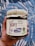 A hand holds a jar labeled 'Imam Bayeldi' with illustrations of eggplants and a pepper. The label emphasizes it is a vegan product, non-GMO, and weighs 16 ounces (454 grams). The jar has a rustic, wrapped lid tied with twine. The background features a blue and white abstract pattern.