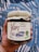 A hand holds a jar labeled 'Imam Bayeldi' with illustrations of eggplants and a pepper. The label emphasizes it is a vegan product, non-GMO, and weighs 16 ounces (454 grams). The jar has a rustic, wrapped lid tied with twine. The background features a blue and white abstract pattern.