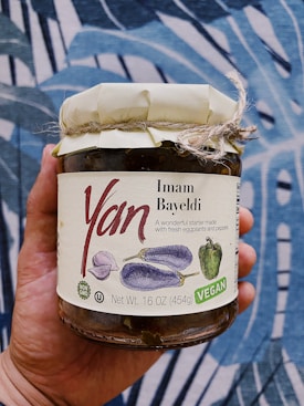 A hand holds a jar labeled 'Imam Bayeldi' with illustrations of eggplants and a pepper. The label emphasizes it is a vegan product, non-GMO, and weighs 16 ounces (454 grams). The jar has a rustic, wrapped lid tied with twine. The background features a blue and white abstract pattern.