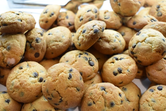 A pile of freshly baked chocolate chip cookies with a golden-brown color. The cookies appear soft and have visible dark chocolate chips scattered throughout. They are arranged closely together, likely on a tray or baking sheet.