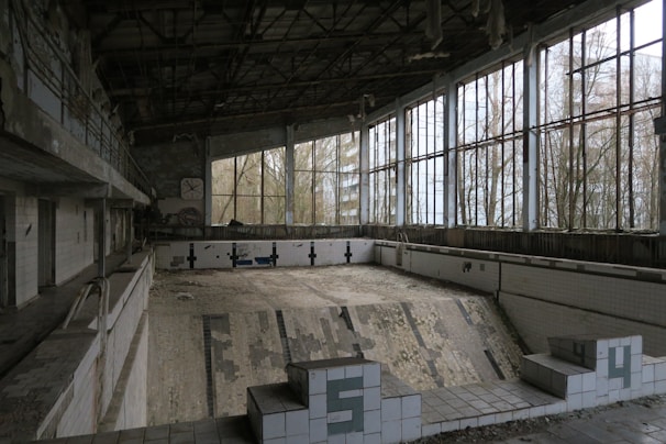 An abandoned indoor swimming pool with cracked tiles on the floor and walls. Large windows with broken glass overlook overgrown trees. The structure is in a state of decay with the ceiling and walls showing signs of disrepair.