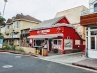 A charming red pizzeria with a striped awning stands on a corner, surrounded by other storefronts. People are gathered near the entrance, adding a lively urban atmosphere. The surrounding buildings offer a mix of architectural styles, and there is lush greenery nearby.