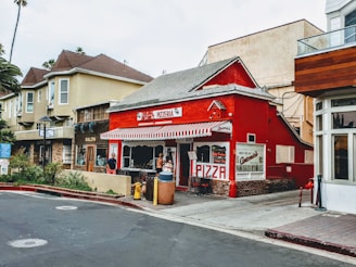 A charming red pizzeria with a striped awning stands on a corner, surrounded by other storefronts. People are gathered near the entrance, adding a lively urban atmosphere. The surrounding buildings offer a mix of architectural styles, and there is lush greenery nearby.