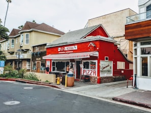 A charming red pizzeria with a striped awning stands on a corner, surrounded by other storefronts. People are gathered near the entrance, adding a lively urban atmosphere. The surrounding buildings offer a mix of architectural styles, and there is lush greenery nearby.