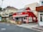 A charming red pizzeria with a striped awning stands on a corner, surrounded by other storefronts. People are gathered near the entrance, adding a lively urban atmosphere. The surrounding buildings offer a mix of architectural styles, and there is lush greenery nearby.