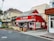 A charming red pizzeria with a striped awning stands on a corner, surrounded by other storefronts. People are gathered near the entrance, adding a lively urban atmosphere. The surrounding buildings offer a mix of architectural styles, and there is lush greenery nearby.