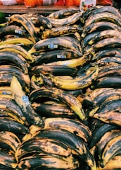 A large pile of ripe plantains with dark spots is displayed at a market stall, each having a blue sticker label. The background features some packaged produce with bright orange and red netting.