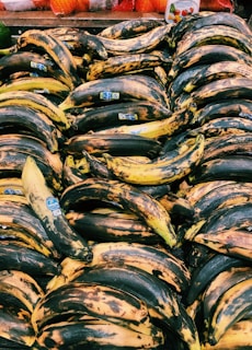 A large pile of ripe plantains with dark spots is displayed at a market stall, each having a blue sticker label. The background features some packaged produce with bright orange and red netting.