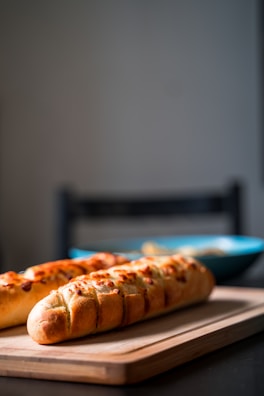 Two baguettes with a crispy crust are placed on a wooden board. The focus is on the bread in the foreground, with a blurred background that includes a blue bowl.