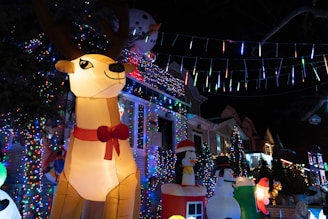 A nighttime scene featuring festive holiday decorations. An inflatable reindeer wearing a red bow is prominently displayed, surrounded by colorful lights. Other inflatable figures include a snowman and a penguin, enhancing the cheerful atmosphere. The houses in the background are adorned with strings of vibrant Christmas lights.
