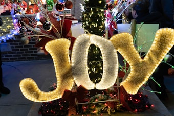 Large illuminated letters spelling 'JOY' are prominently displayed, adorned with lights and surrounded by festive decorations including ribbons and ornaments. In the background, there are more holiday lights and decorations on a brick surface. The scene has a warm and celebratory atmosphere.