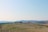 Rolling hills stretch across the landscape with patches of green fields and dry, rocky soil in the foreground. A winding path cuts through the hills, and a cluster of trees and a structure are seen in the distance under a clear blue sky.