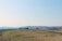 Rolling hills stretch across the landscape with patches of green fields and dry, rocky soil in the foreground. A winding path cuts through the hills, and a cluster of trees and a structure are seen in the distance under a clear blue sky.