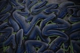 Abstract graffiti art featuring intertwined humanoid figures. The figures are dark blue with metallic, robotic details and are outlined with neon green accents. They appear to be stacked or layered in a fluid, wave-like pattern.