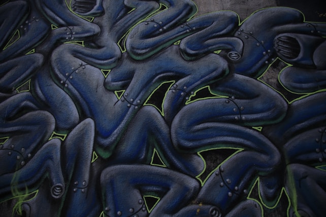 Abstract graffiti art featuring intertwined humanoid figures. The figures are dark blue with metallic, robotic details and are outlined with neon green accents. They appear to be stacked or layered in a fluid, wave-like pattern.